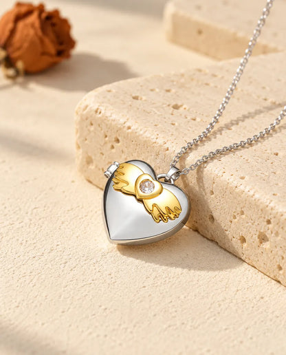 Personalised angel wings heart photo locket necklace in silver tone with gold tone angel wings, resting on a travertine block