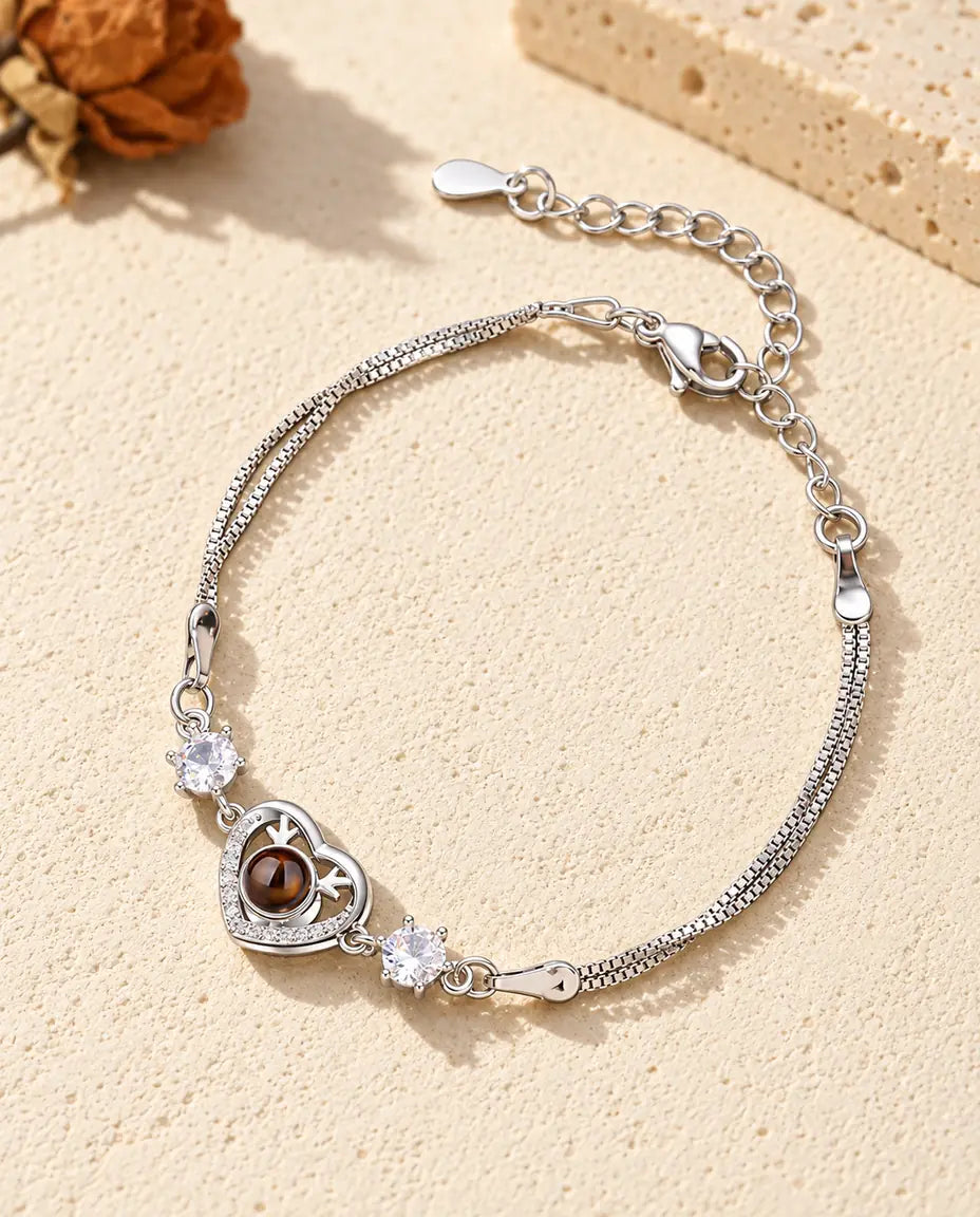 Personalised photo projection bracelet with a silver tone antler heart charm and two cubic zirconia on a double chain, lying on cream plaster