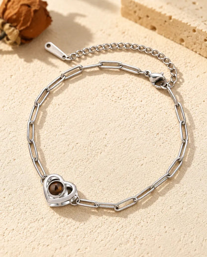 Personalised photo projection bracelet in silver tone with a double heart charm on a paperclip chain, lying on cream plaster
