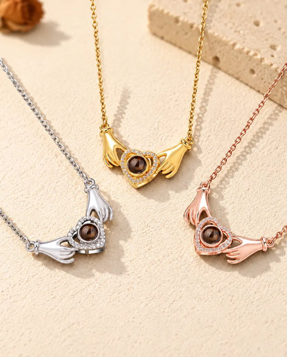 Three colours of the hands holding heart photo projection necklace in a row on cream plaster