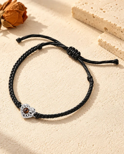 Personalised photo projection bracelet with a silver tone openwork heart charm on a black thin braided cord, lying on cream plaster