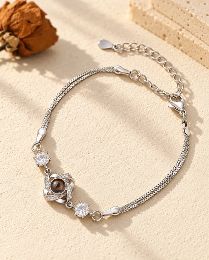 Personalised love knot photo projection bracelet in silver tone with two cubic zirconia on a double box chain, lying on cream plaster