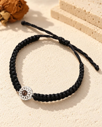 Personalised photo projection bracelet with a silver tone openwork round charm on a black braided cord, lying on cream plaster